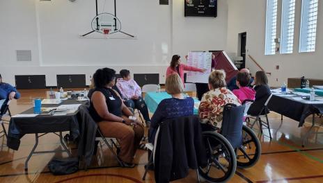 A group of people are gathered around a table in a gymnasium, watching a presenter point to a flipchart